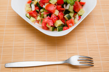 Fresh salad of finely chopped cucumbers, tomatoes and onions with sunflower oil in a white plate and a fork on a wooden table. The concept of healthy nutrition and eco-friendly, vegetarian diet.