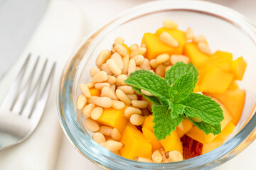 Fruit salad close up. Sliced mango, peach, pine nuts, and mint leaf in a glass bowl on white background, view from above.
