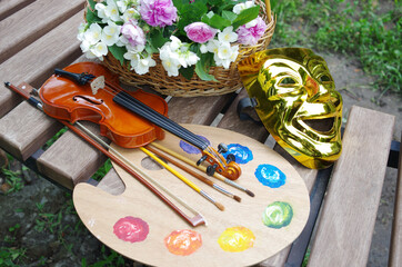 Attributes of the arts. Art palette, brushes, violin, bow, theater mask, a bouquet of roses and jasmine on a garden bench.