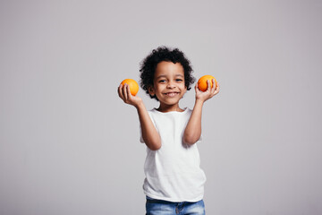 Curly African American boy in a white T-shirt holds two oranges in his hands and smiles on a gray...