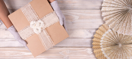 Safe celebration party. Hands holding white gift ribbon box with rubber gloves, over wooden background. Top view