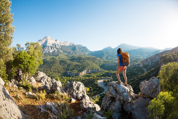 A woman with a backpack stands on top of a mountain and admires the beauty of a mountain valley.