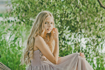 Obraz premium Portrait of pretty caucasian girl sitting near the lake on sunny day. Lovable long-haired blonde girl posing on a nature background.