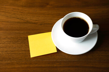 A white cup of hot Americano coffee with saucer and blank orange stick note paper on wooden table.Orange stick note paper with copy space for text.Top view.