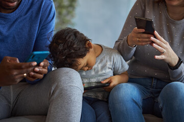 Concentrated members of family sit together on one sofa. Each of them has phone in hands. They look...