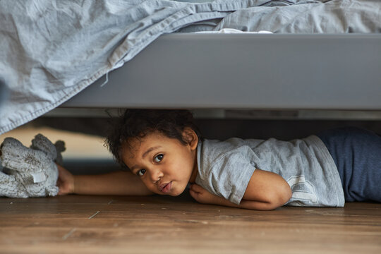Family at home concept, boy playing hide and seek. Cute afro boy peeking out from under the bed.
