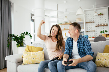 Loving couple playing video games with joysticks in living room. Husband and wife are playing video games at home.	