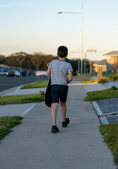 Boy from behind walking home from skate park with his skateboard under his arm © Em Neems Photography
