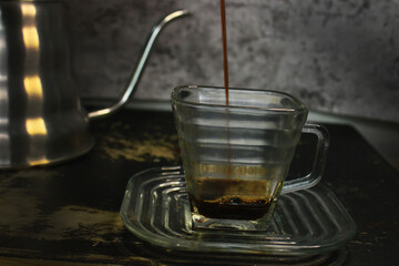 the process of pouring espresso produced mocha pot into a square-shaped coffee cup before mixed with milk, on the table with a swan neck kettle on the back of the glass