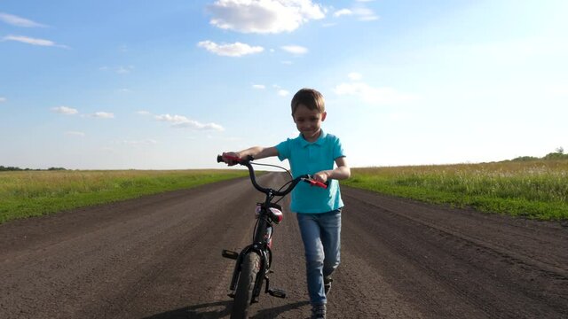 A happy smiling boy is walking along a country road, holding a Bicycle. A child rides a bike. Outdoor activity.