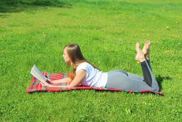 A beautiful young white girl in a white T-shirt and with long hair lies on a red plaid, on green grass, on the lawn and read book