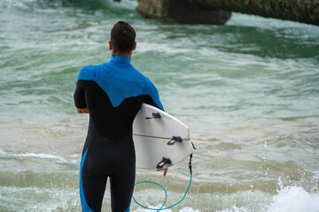 Male surfer wearing black wetsuit and holding his surfboard under his arm, walking into the surf,...