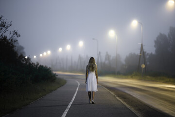 One young alone abandoned woman in white dress slowly walking on long sidewalk under street lights...