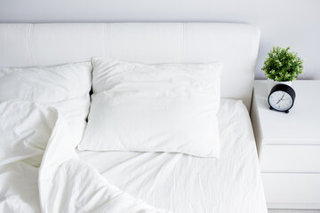 top view of empty unmade bed with white blanket and two pillows, bedside table with alarm clock and...