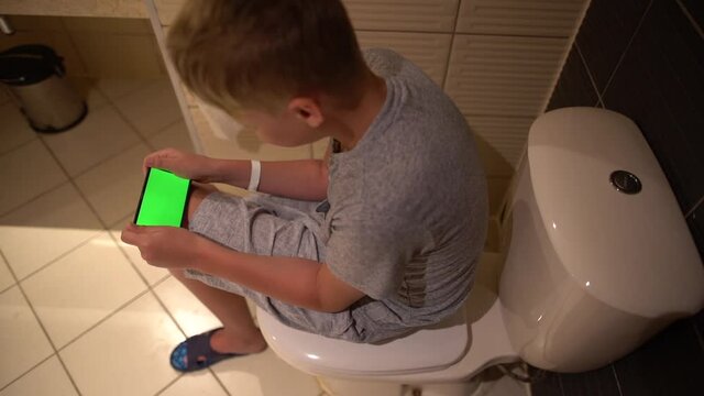 Closeup top view of young kid's hands holding modern mobile smartphone with empty green screen while hiding from parents sitting on toilet bowl closed with cover.