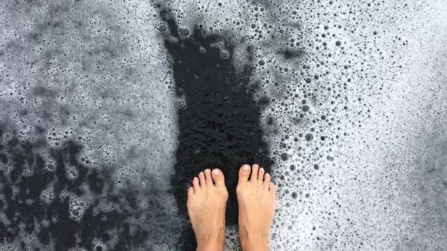 Women's feet standing in black sand in Hawaii. A wave sweeps over her feet and leaves the frame. Slow-motion