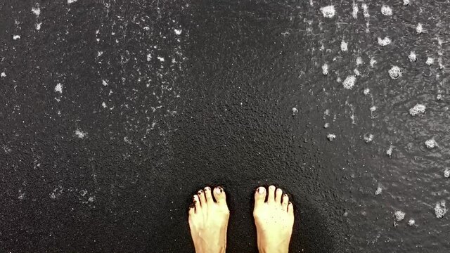 Women's feet standing in black sand in Hawaii. A wave sweeps over her feet and leaves the frame.