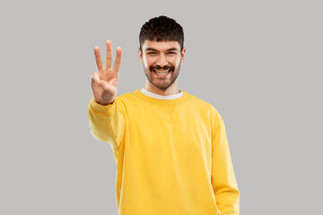 count and people concept - happy smiling young man in yellow sweatshirt showing three fingers over grey background