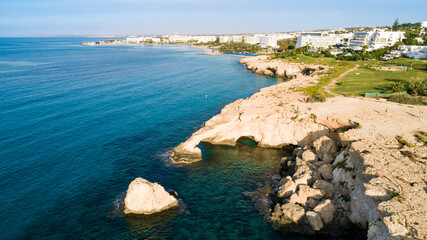 Aerial bird's eye view of coastline landmark ‘Love bridge’ and sea caves at Cavo Greco, Ayia Napa, Famagusta, Cyprus from above. A tourist attraction cliff rock natural formation arch in Ammochostos.
