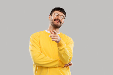 people concept - smiling young man in glasses and yellow sweatshirt pointing finger to camera over grey background