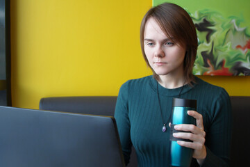 A girl with a calm expression on her face sits at a laptop at a table in a cafe. He is looking at a computer monitor, holding a reusable eco-friendly coffee cup in his hand