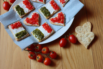 Salad cookies in shape of red heart and italian flag on a plate on wooden table. Snack with tradional  italian ingredient: pesto, cheese and red tomatoes.