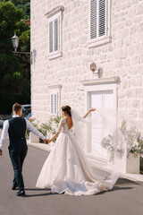 A wedding couple walks along the promenade against the background of a white old house, the bride waves a long veil, the groom holds her hand. Fine-art wedding photo in Montenegro, Perast.
