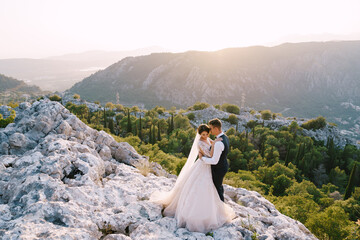 A wedding couple stands on top of a mountain with panoramic views of the Bay of Kotor, at sunset. The groom gently hugs the bride. Fine-art destination wedding photo in Montenegro, Mount Lovchen.