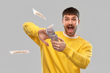 people concept - happy young man in yellow sweatshirt throwing money into air over grey background
