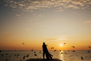 Couple hugging in front of sunset at sea. Watching the sunset with lovers. Kissing couples silhouette. Silhouette of romantic couple. Orange sky with flying seagulls. Copy space.
