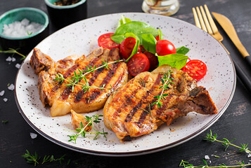 Grilled pork steaks and salad with tomatoes in plate on dark background.