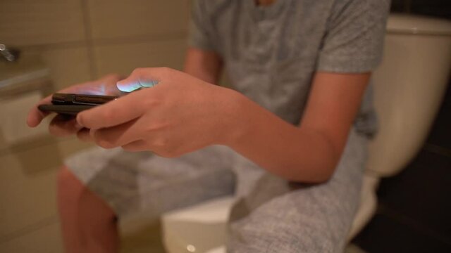 Closeup view of young kid's hands holding modern mobile smartphone to play online video games while hiding from parents sitting on toilet closed with cover bowl. Mother takes away phone and scolds boy