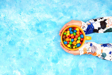 Female hands in colorful clothes holding a big mug of multicolored chocolate candies against a bright blue background. Top view, copy space.
