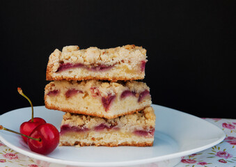 Portions of homemade cherry pie with crumble on white plate and flowered tablecloth, with black background