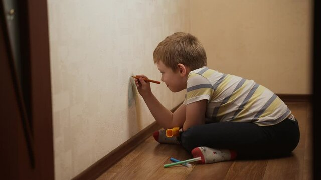 Caucasian child, a boy sitting near the wall licks his lip with his tongue and draws on the wallpaper with a felt-tip pen, marker