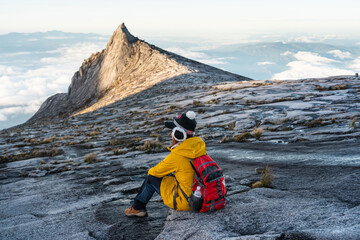 Young woman trekker with backpack sitting in front of South peak of Kinabalu mountain massif,...