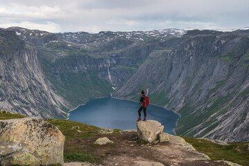 Asian woman traveller with backpack standing on rock and enjoying landscape of mountains and lake in Trolltunga mountain cliff trail, Odda town, Norway, Scandinavia