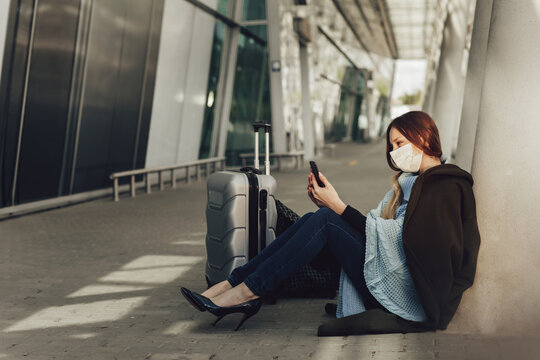 Young woman in medical mask sits near luggage in airport. Woman waiting for flight, using a cell phone and looking at camera. Girl trying to fly out of the country during quarantine. Travel concept