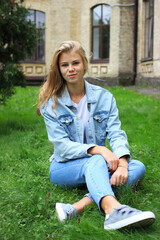 Obraz premium A very charming blonde girl in jeans sits on the grass near the university and is resting.