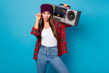 Photo of good mood pretty lady spring day weather rejoicing walking street hold tape recorder shoulder sticking tongue wear casual hat plaid shirt jeans shoes isolated blue color background