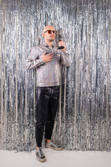 Picture of beautiful caucasian man without hair with black sunglasses in a shiny shirt, black leather pants and grey sneakers sings in a small silver microphone isolated on silver glitter background