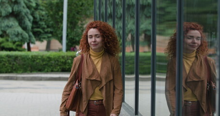 Fototapeta premium Attractive young red-haired curly woman with a bag on her shoulder walks near the glass wall looking at the camera