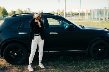 Beautiful young caucasian woman in white jeans, white trainers and black jacket poses for the camera with old black briken car outside