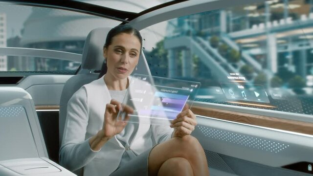 Attractive Senior Female Reading News on a Futuristic Transparent Tablet Computer with Augmented Reality Interface while Sitting on a Backseat of Autonomous Car. Self-Driving Van Rides on Public Road.