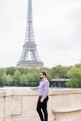Handsome bearded young man in glasses and casual checkered shirt enjoying wonderful view of Paris and Seine river, standing on the bridge and looking away. Eiffel tower on the background