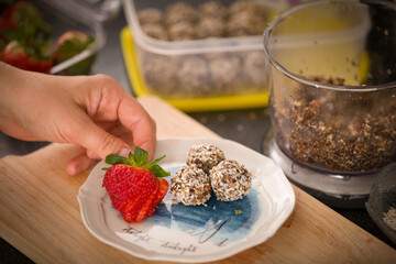 Top view on a plate with delicious homemade protein balls and ripe strawberries in the girl's hand. Healthy sweets. Organic raw treats from nuts, coconut, dates and cocoa