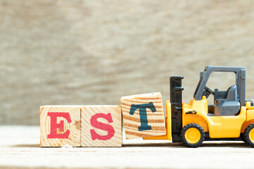 Toy forklift hold letter block t to complete word EST (abbreviation of established, estimated, eastern time zone, expressed sequence tag) on wood background