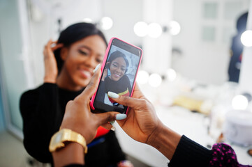 African American woman applying make-up by make-up artist at beauty saloon. Artist make photo on...