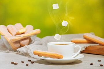White cup of hot aromatic coffee with steam and falling sugar cubes. Traditional italian savoiardi biscuits or ladyfingers cookies on a plate and in a glass bowl. Selective focus