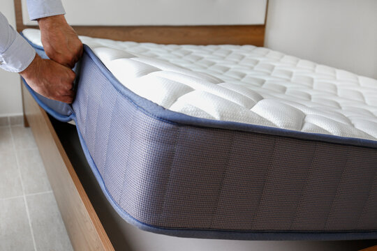 New Bed Delivery And Assembly Service Concept. Cropped Shot Of Male Worker's Hands In Process Of Laying The Orthopedic Foam Mattress On Carcass In Customer's Bedroom. Close Up, Copy Space, Background.